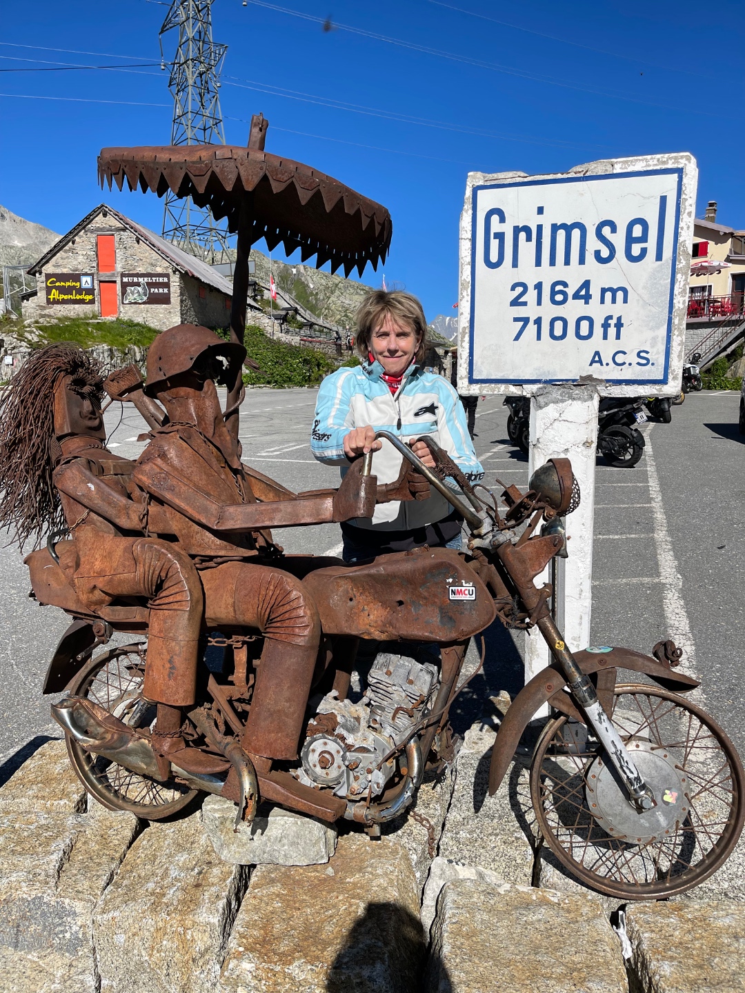 Kathy posing with rusty iron motorcycle sculpture at Grimsel Pass 2164m 7100ft sign, Switzerland